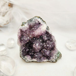 A raw amethyst crystal cluster with purple and pink druzy cavities, resting on a white surface with clear crystal pebbles and a soft white fabric backdrop.